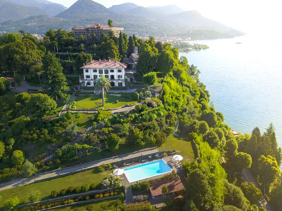 A large real estate villa with a pool sits on a lush hillside overlooking a lake, surrounded by greenery and mountains in the background.