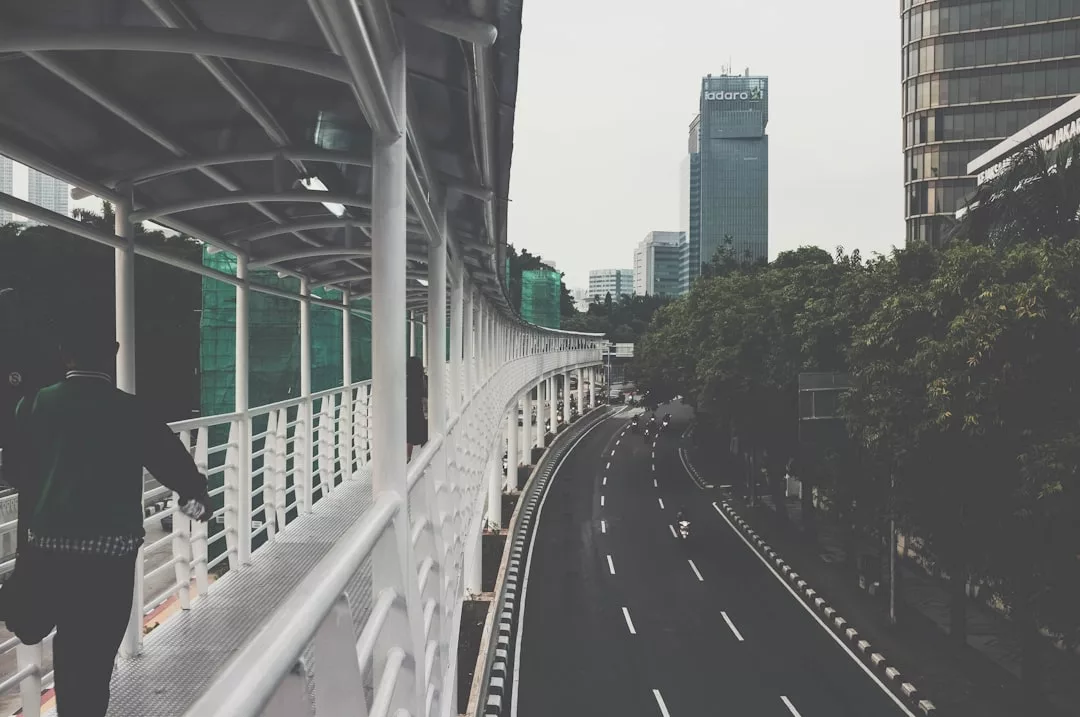 A man walks on a covered pedestrian bridge above an empty multilane road in an urban area, surrounded by tall buildings, trees, and vibrant real estate developments.