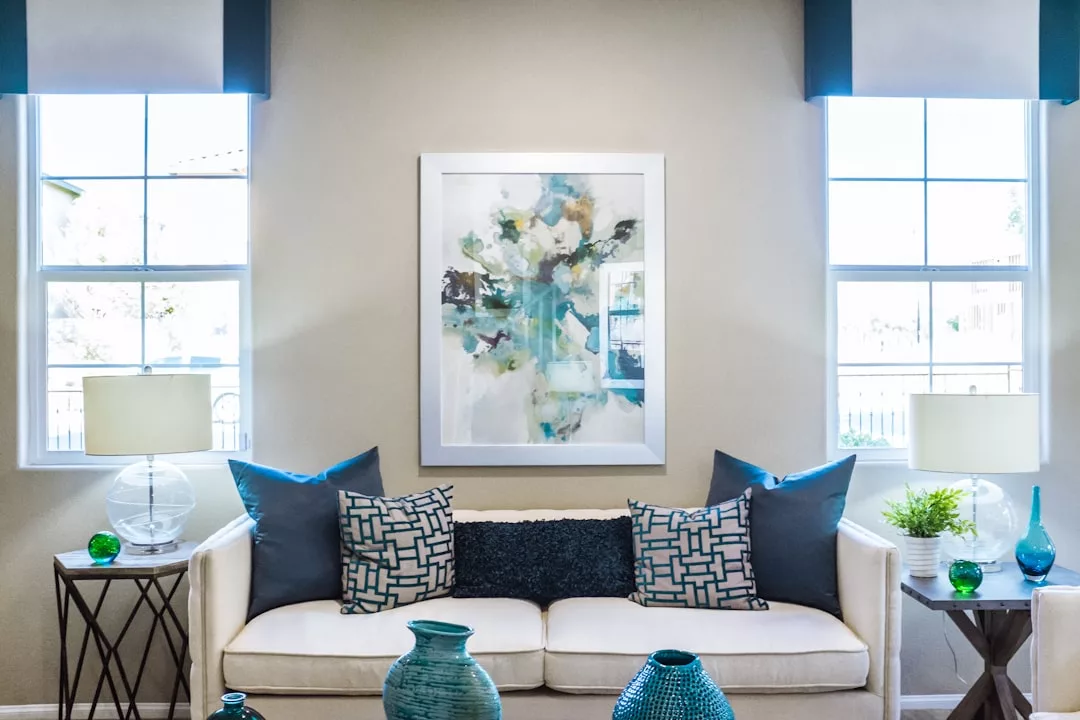 A modern living room ideal for real estate showcases a white sofa, blue and gray accent pillows, side tables with lamps, abstract wall art, and decorative vases. Two windows fill the space with natural light.