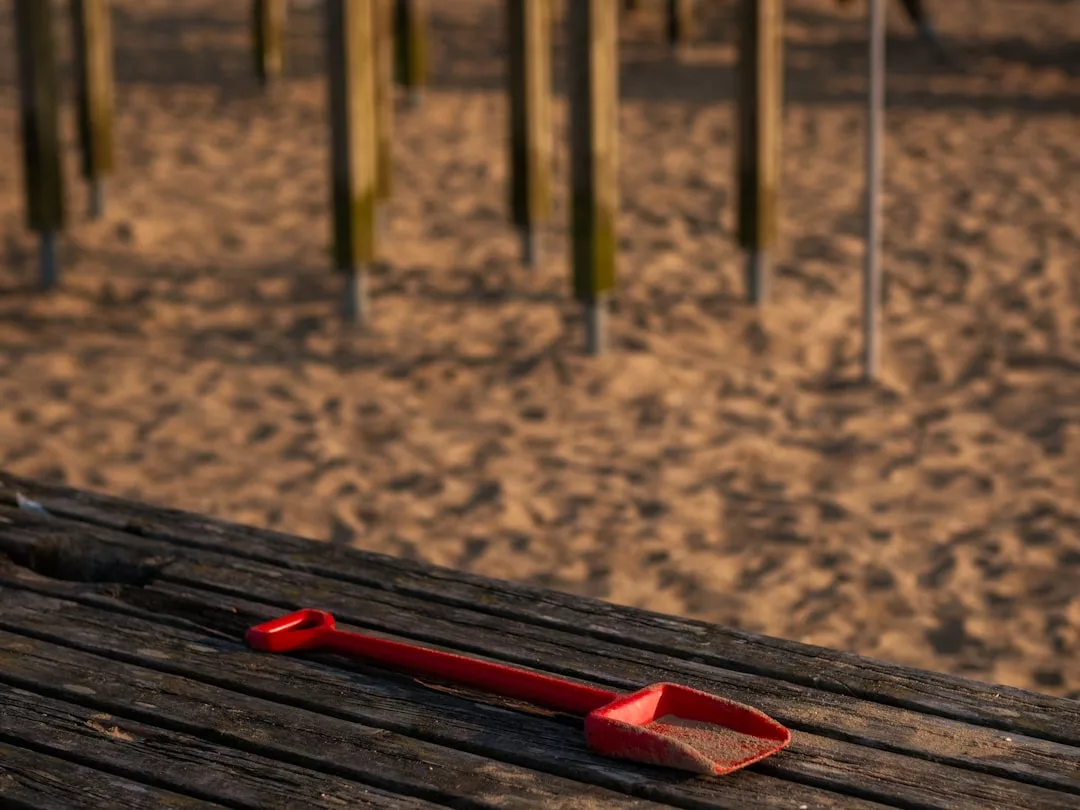 A red plastic shovel lies on a wooden surface with sand, evoking the playful beginnings of real estate dreams, while vertical wooden posts rise in the blurred background.