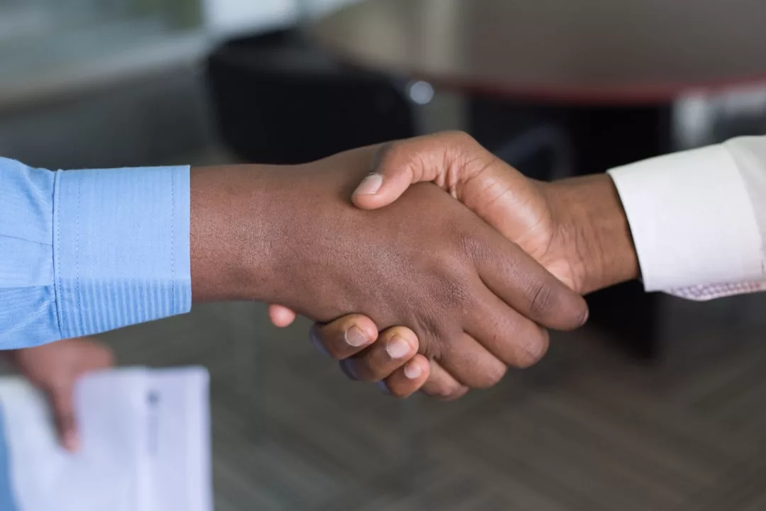 Two people in business attire shaking hands over a table, with one person holding a real estate document in their left hand.