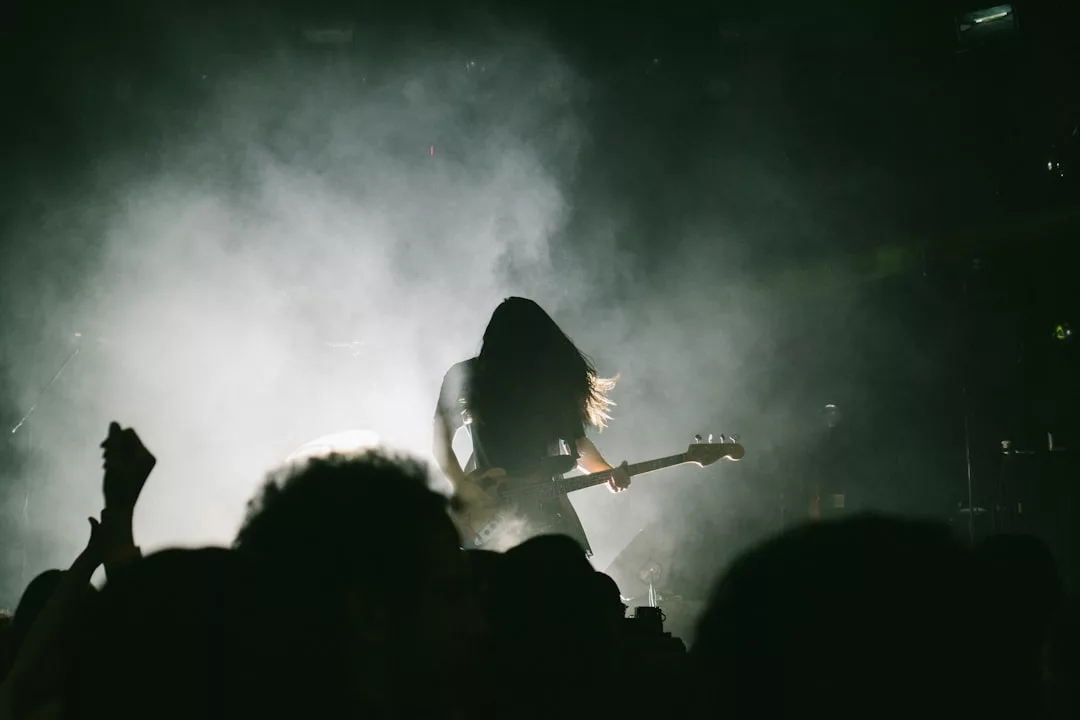 A musician with long hair plays an electric guitar on stage, surrounded by smoke and dim lighting, as an audience—and even a few real estate enthusiasts—watch in the foreground.