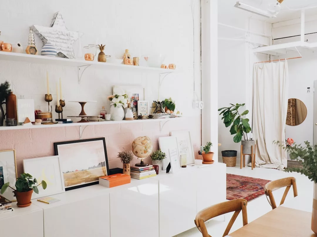 A bright, modern room with white walls, wooden chairs, plants, art, and decorative items on shelves—a perfect showcase for selling luxury properties in Oslo. A stylish rug and curtain complete the sophisticated atmosphere.