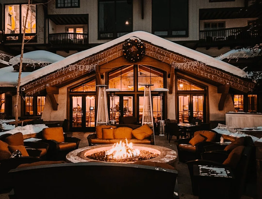 Outdoor seating area with cushioned chairs arranged around a fire pit, set before a snow-covered building adorned with lights and a wreath—perfect for enjoying winter evenings and enhancing the curb appeal of your real estate at night.
