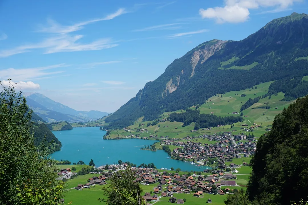 A turquoise lake borders a small village and green hills, with forested mountains rising in the background—an idyllic setting perfect for real estate opportunities under a blue sky with scattered clouds.