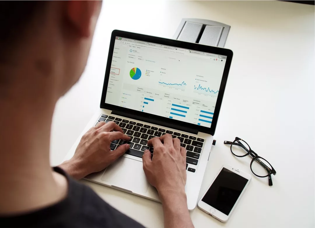 Person typing on a laptop displaying real estate data analytics charts; eyeglasses and a smartphone are placed on the desk nearby.
