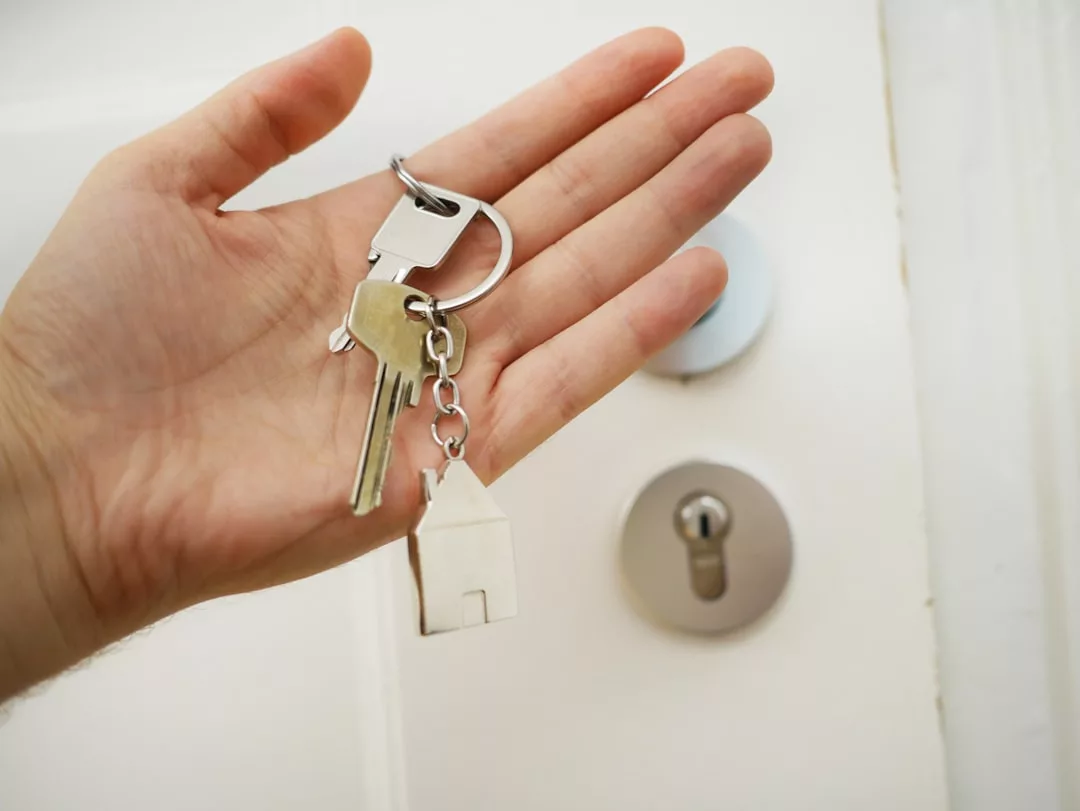 A hand holds a keyring with two keys and a small house-shaped keychain in front of a white door with a lock, symbolizing the excitement of real estate transactions.