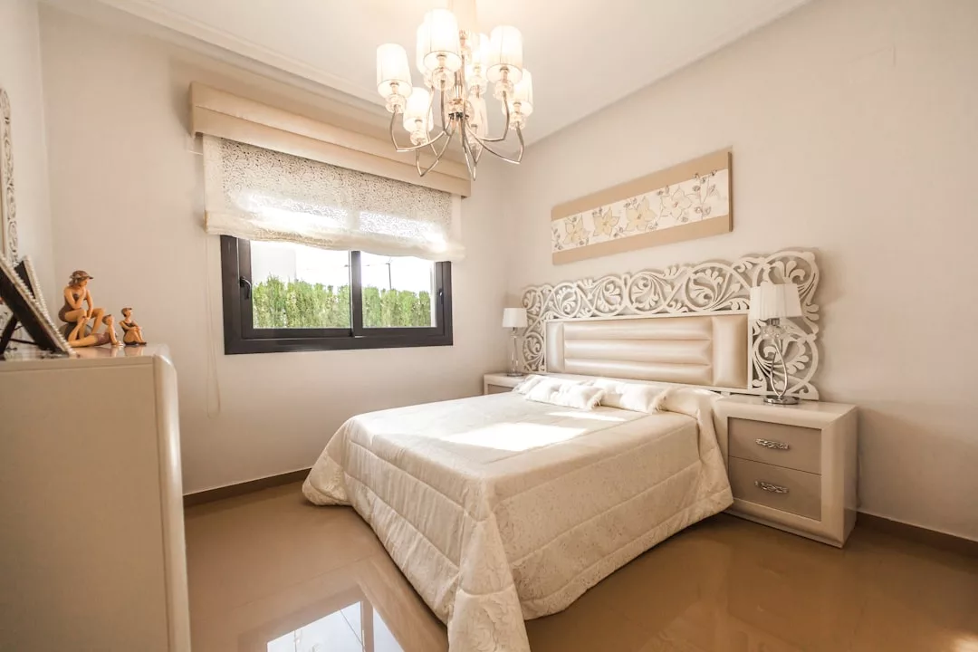 A bright Real Estate bedroom featuring a double bed with an ornate headboard, two nightstands, a dresser adorned with figurines, a window with blinds, and an elegant chandelier.