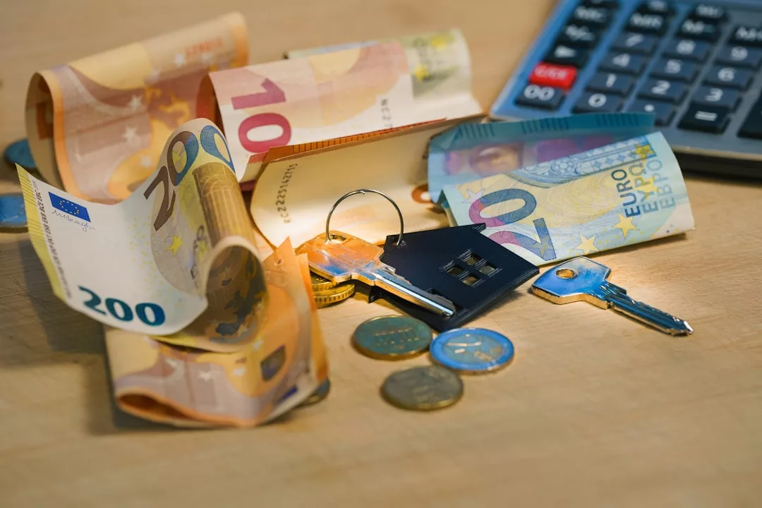 Euro banknotes, coins, two keys, and a keychain shaped like a house are spread on a table next to a calculator, symbolizing real estate finances and property investment.