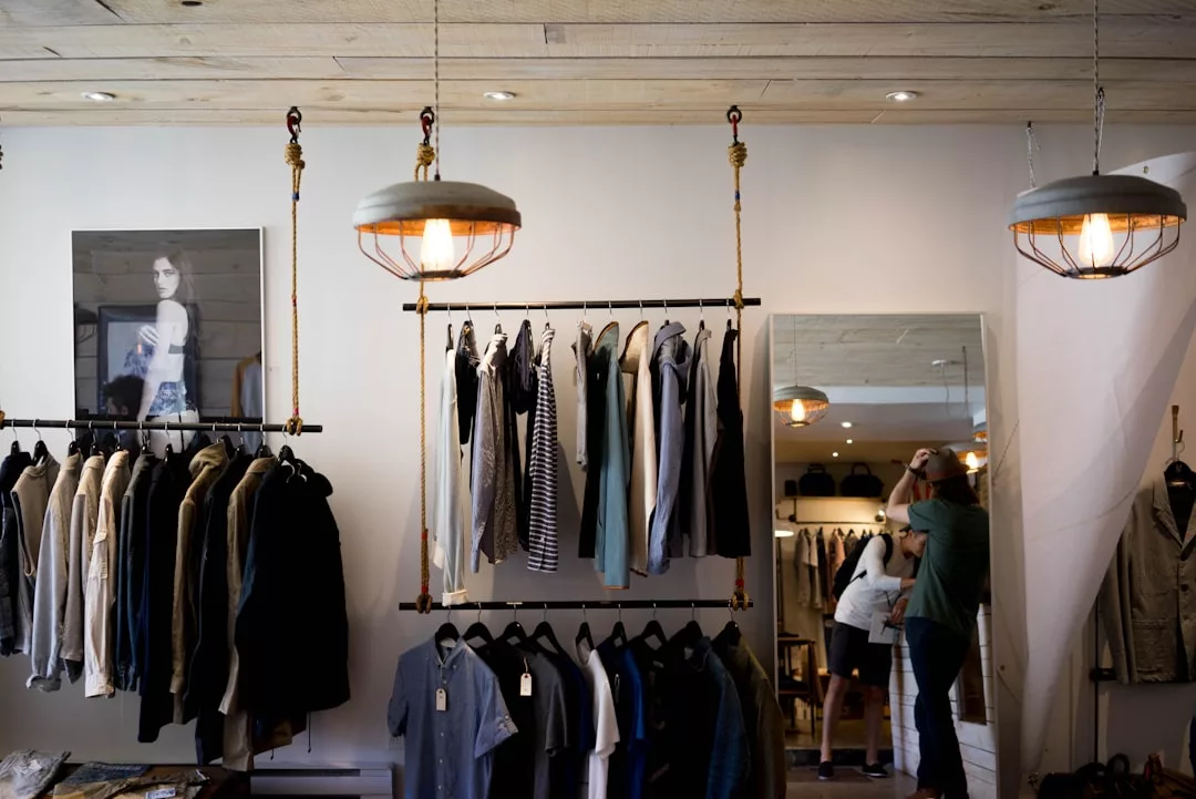 Clothing racks with assorted garments in a modern boutique, set within a stylish real estate space, with two people looking at items near a mirror in the background.