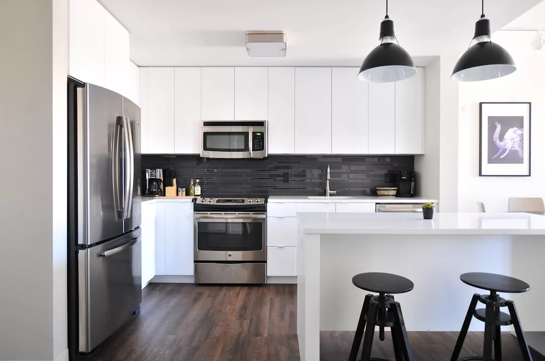 Modern kitchen with white cabinets, stainless steel appliances, a black backsplash, center island with two black stools, and two black pendant lights—a stylish space perfect for your property in Oslo.