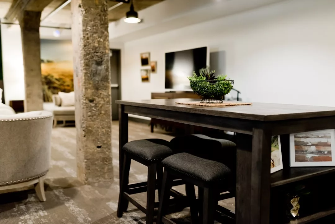 A modern Real Estate listing showcases a dark wooden table with two cushioned stools, a potted plant centerpiece, exposed concrete columns, and a TV on a stand against a crisp white wall.