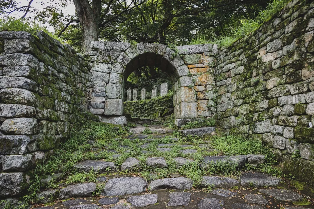 Stone pathway leading to an arched stone gate with moss-covered walls and trees in the background—an enchanting real estate opportunity blending natural beauty with timeless charm.