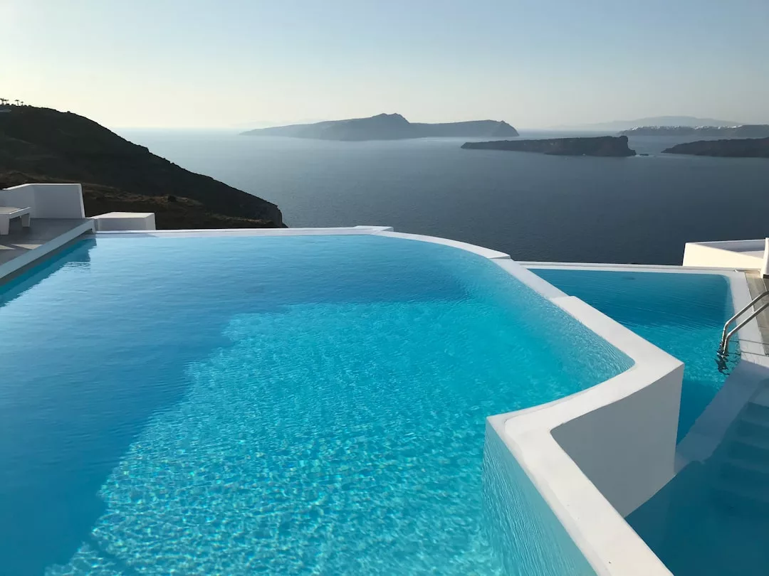 Infinity pool with clear blue water overlooking a calm sea and distant islands, surrounded by white modern architecture under a clear sky—an exceptional real estate opportunity for those seeking luxury and tranquility.