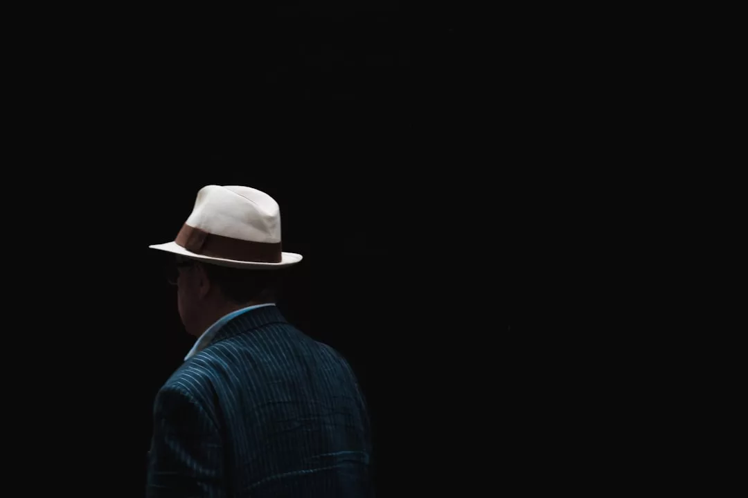 A person wearing a white fedora and a striped jacket is shown from behind, standing against a dark, black background—evoking the poised confidence of a real estate professional ready to make bold moves.