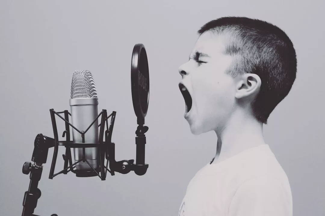 A child faces a large microphone and pop filter, mouth wide open as if singing or shouting, against a plain background—capturing enthusiasm reminiscent of an agent announcing exciting real estate opportunities.
