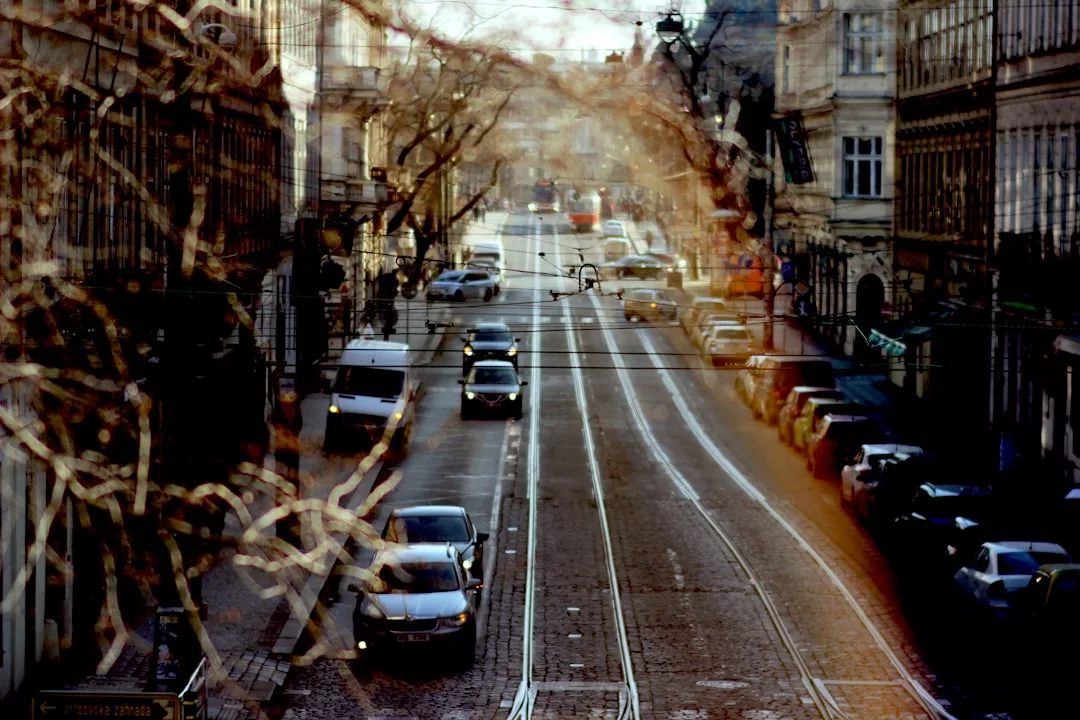 A city street with tram tracks, parked cars, and moving vehicles captures urban Real Estate life, overlaid with a second, blurry layer of branches and leaves.