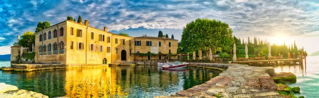 Large yellow villa surrounded by water, boats docked nearby, stone pathway, and lush trees under a partly cloudy sky at sunset—an exquisite real estate opportunity.