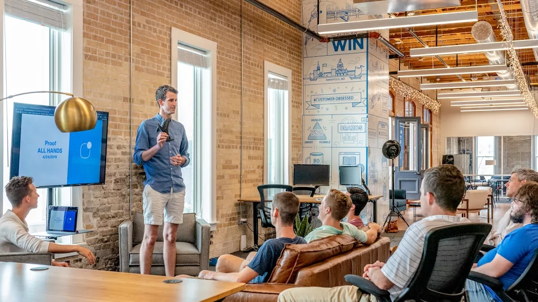 A man stands and speaks to a group of people seated in a modern office space, presenting key Real Estate trends displayed on a screen next to him.