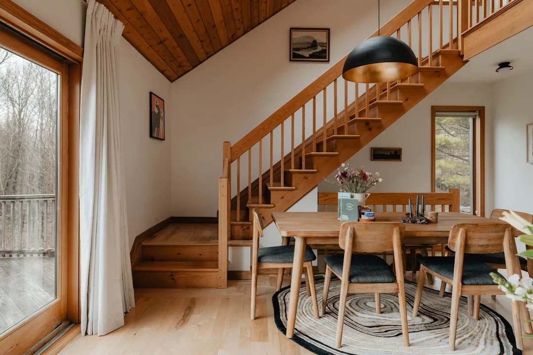 A dining area with a wooden table and chairs sits beside a staircase with wood railings; large windows and framed art enhance the inviting real estate setting.