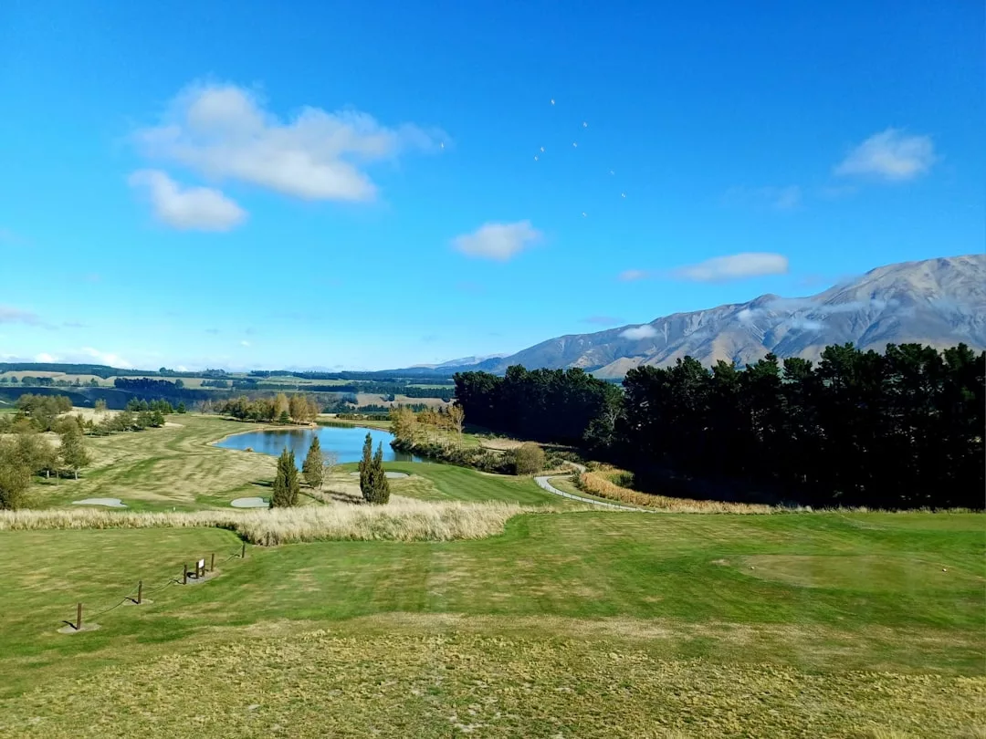 A golf course with a pond, trees, and mountains in the background under a blue sky with scattered clouds—an ideal setting for prime real estate.