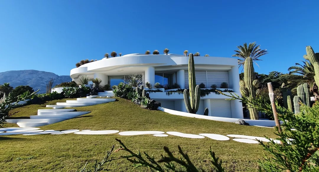 A modern white house with curved lines sits on a grassy hill, surrounded by cacti, palm trees, and landscaped greenery under a clear blue sky—a standout in contemporary real estate.