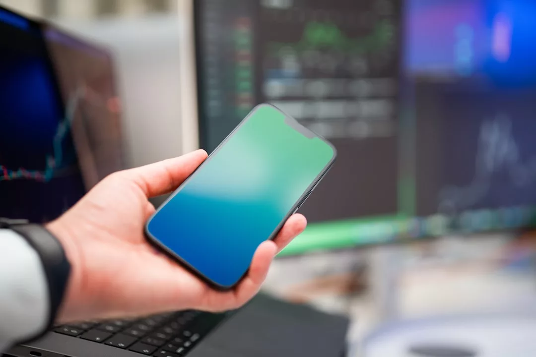 A person holds a smartphone with a blank screen in front of computer monitors displaying real estate financial charts and data.