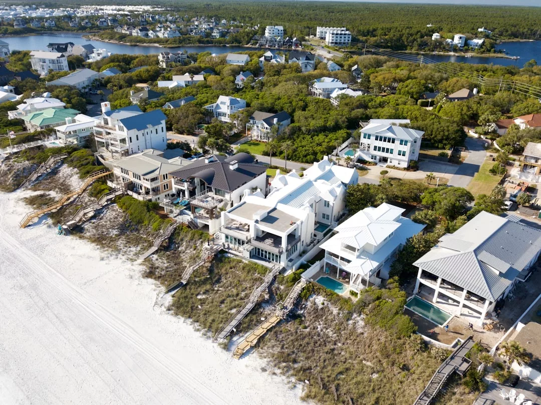 Aerial view of beachfront real estate with private walkways leading to a sandy beach, surrounded by lush greenery and a residential neighborhood in the background.