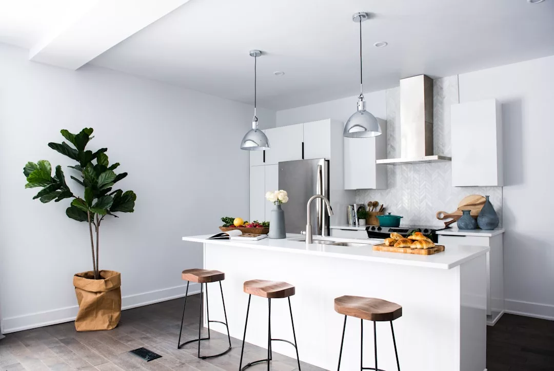 Modern real estate kitchen with white cabinets, stainless steel appliances, an island with three wooden stools, potted plant, pendant lights, and assorted food items on the counter.