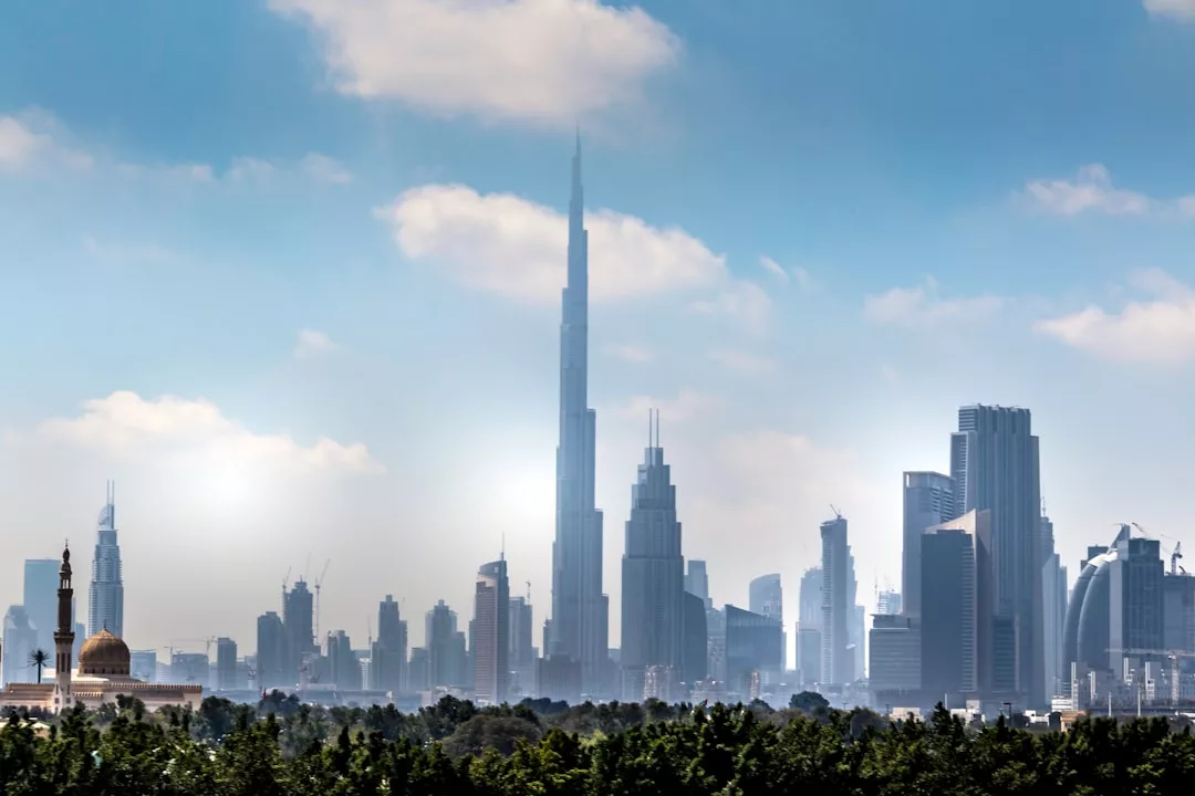 A city skyline with modern skyscrapers under a partly cloudy sky, prominently featuring the Burj Khalifa in the center. Lush trees in the foreground highlight a vibrant real estate landscape.