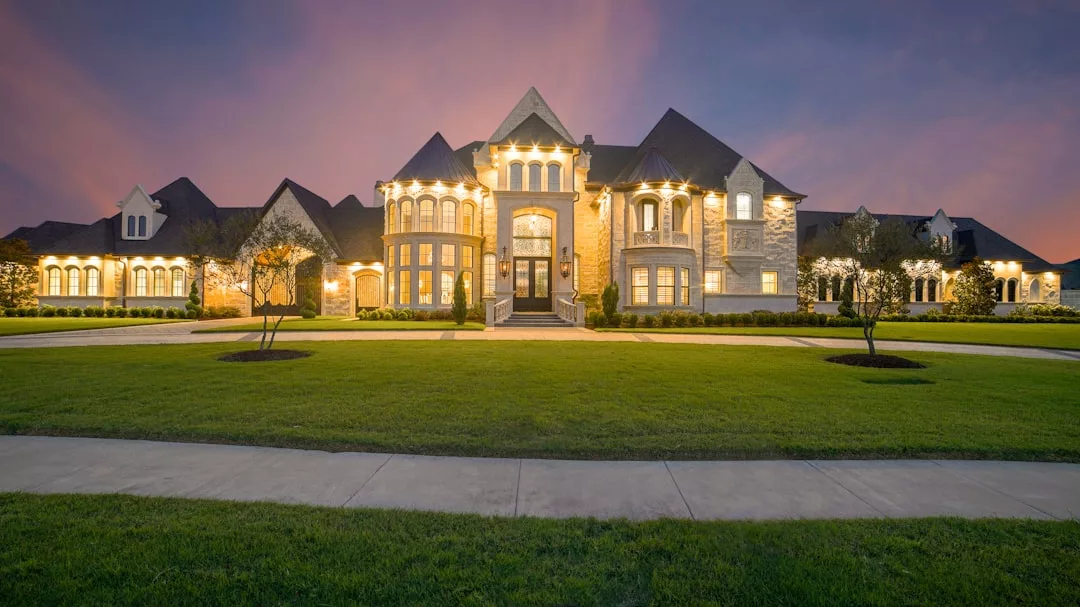 Large, well-lit real estate mansion with multiple gables, stone facade, and manicured lawn, photographed at dusk under a purple sky.