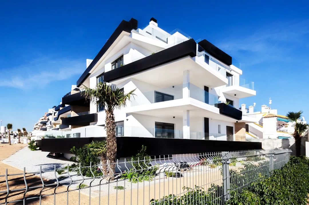 Modern white apartment building with black accents, multiple balconies, and large windows; palm trees and a metal fence are in the foreground under a clear blue sky—an impressive piece of real estate.