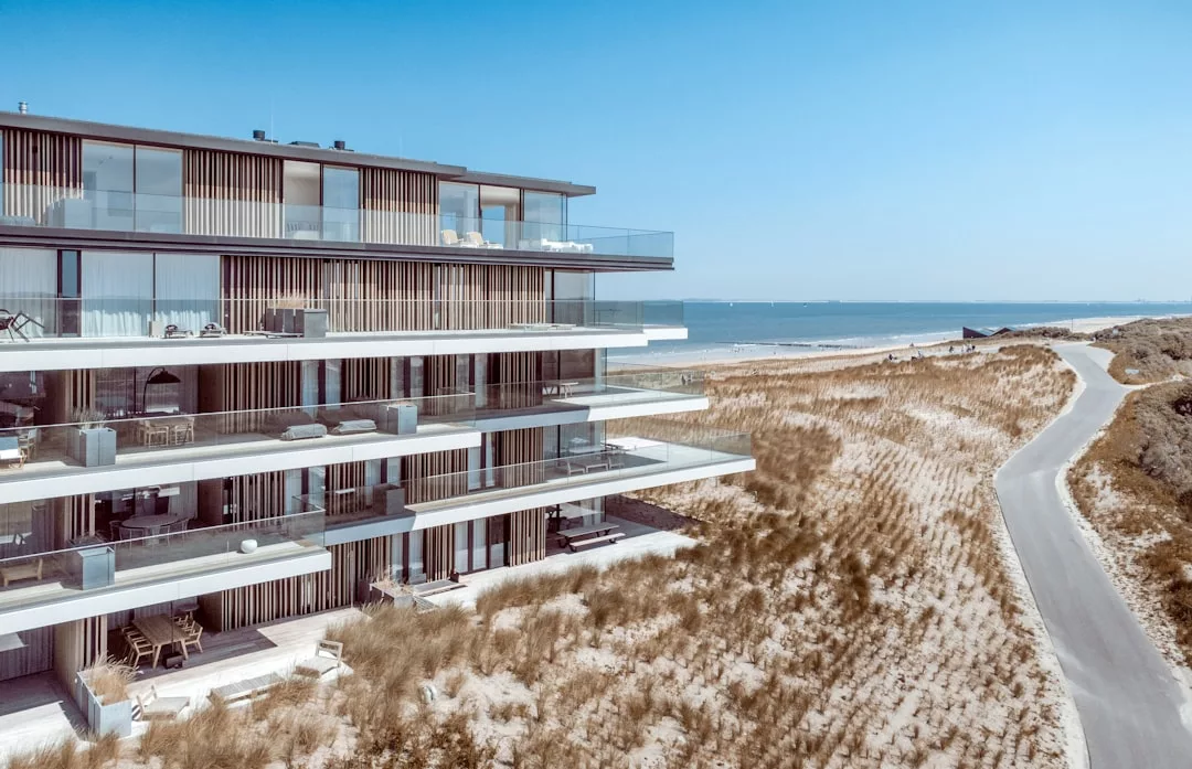 Modern beachfront real estate apartment building with glass balconies, surrounded by sand dunes and a narrow road, overlooking the sea under a clear blue sky.