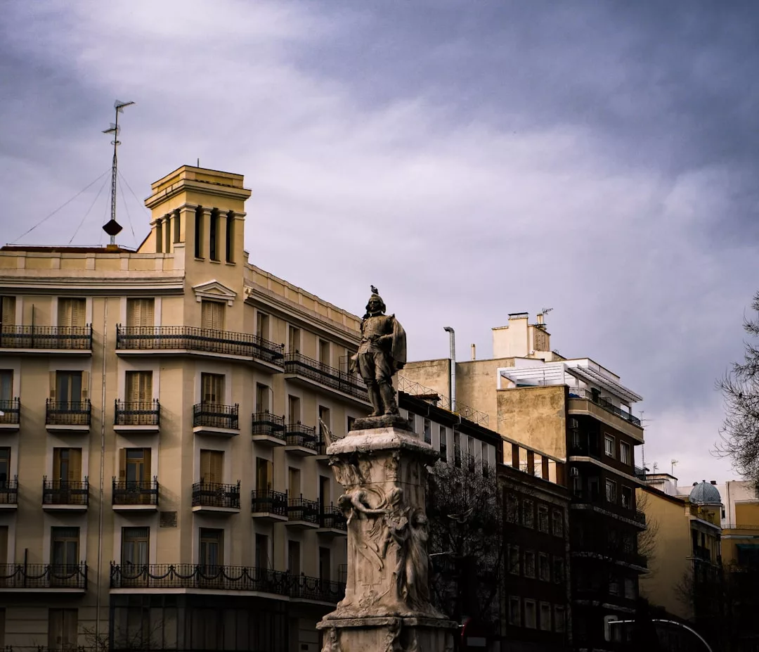 A stone statue stands on a decorated pedestal in front of multi-story yellow and brown real estate buildings under a cloudy sky.