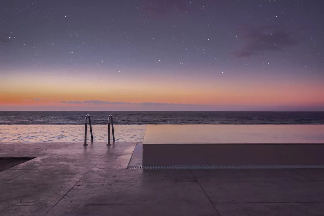 An infinity pool overlooks the ocean at sunset, with a clear sky and stars above the horizon—an exquisite real estate opportunity for those seeking luxury living by the sea.