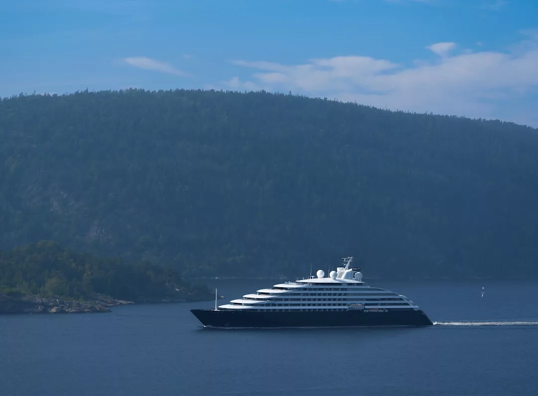 A large, modern cruise ship sails on calm water past a forested hillside, its sleek lines and amenities rivaling floating real estate, beneath a partly cloudy sky.