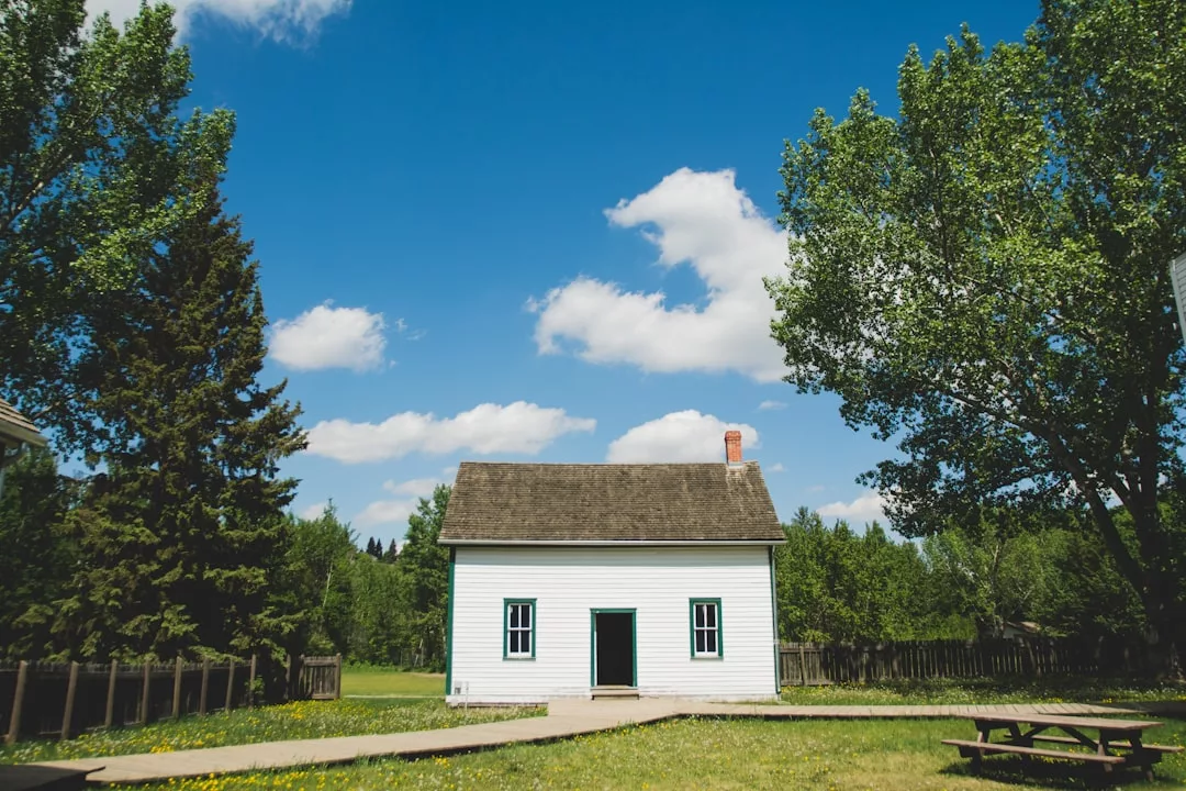 A small white house with blue trim, perfect for real estate seekers, stands surrounded by trees under a blue sky with scattered clouds. A wooden picnic table sits invitingly in the foreground.