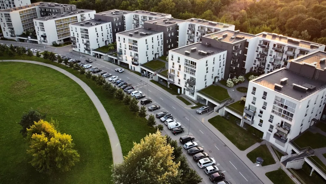 Aerial view of modern apartment buildings in one of the popular neighborhoods Oslo offers, next to a curved park and parked cars along the street, with dense trees in the background.
