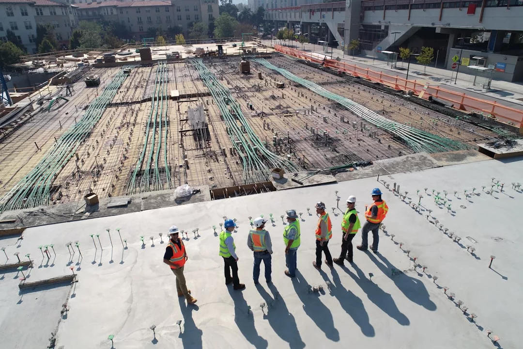 Six construction workers in safety gear stand on a concrete slab, discussing plans at a large real estate building site with materials and unfinished sections visible.