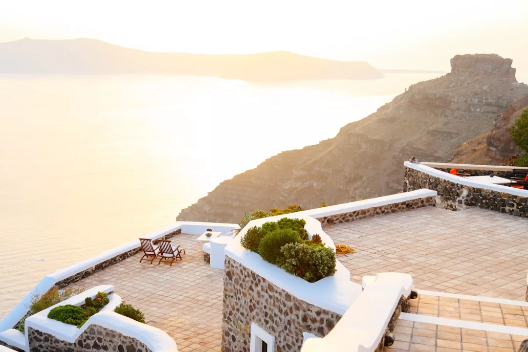 Terrace with white walls and stone tiles overlooks the sea and rocky hills at sunset; perfect for relaxing, this Real Estate gem features two lounge chairs and lush plants on the terrace.