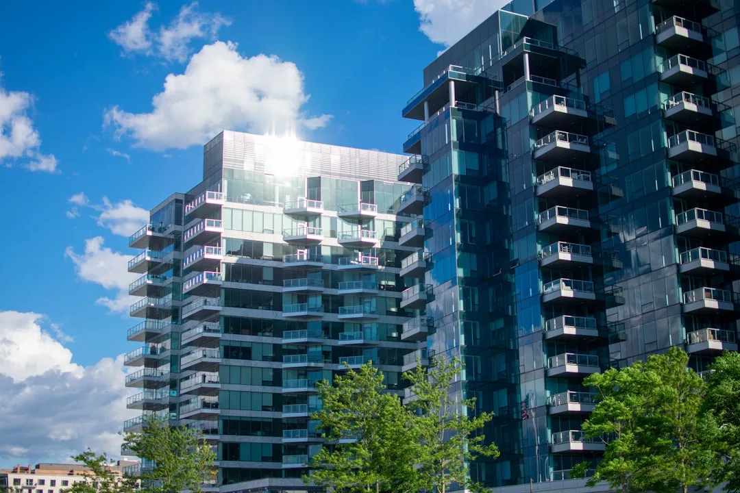 Modern glass apartment buildings with balconies under a blue sky in Oslo, sunlight reflecting off the windows, and green trees in the foreground.