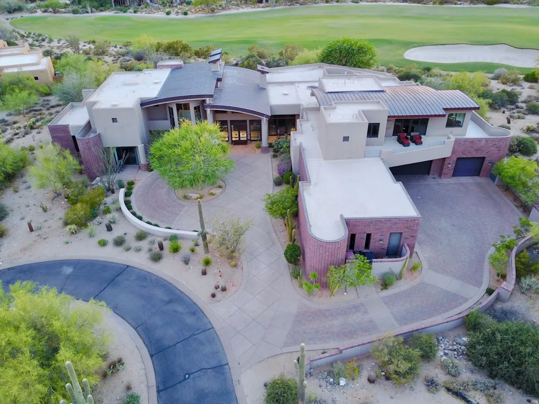 Aerial view of a modern, multi-level real estate property with flat roofs, a circular driveway, and desert landscaping, adjacent to a golf course.