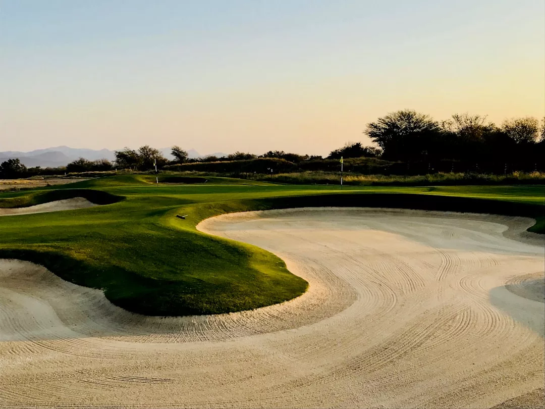 A golf course with manicured greens, sand bunkers, and flags at dusk, surrounded by trees and distant mountains—an ideal landscape for luxury real estate.