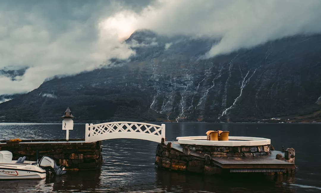 A small dock with a white bridge and circular seating area overlooks a lake, offering stunning waterfront views of misty mountains and waterfalls—one of the top locations for properties in Oslo.