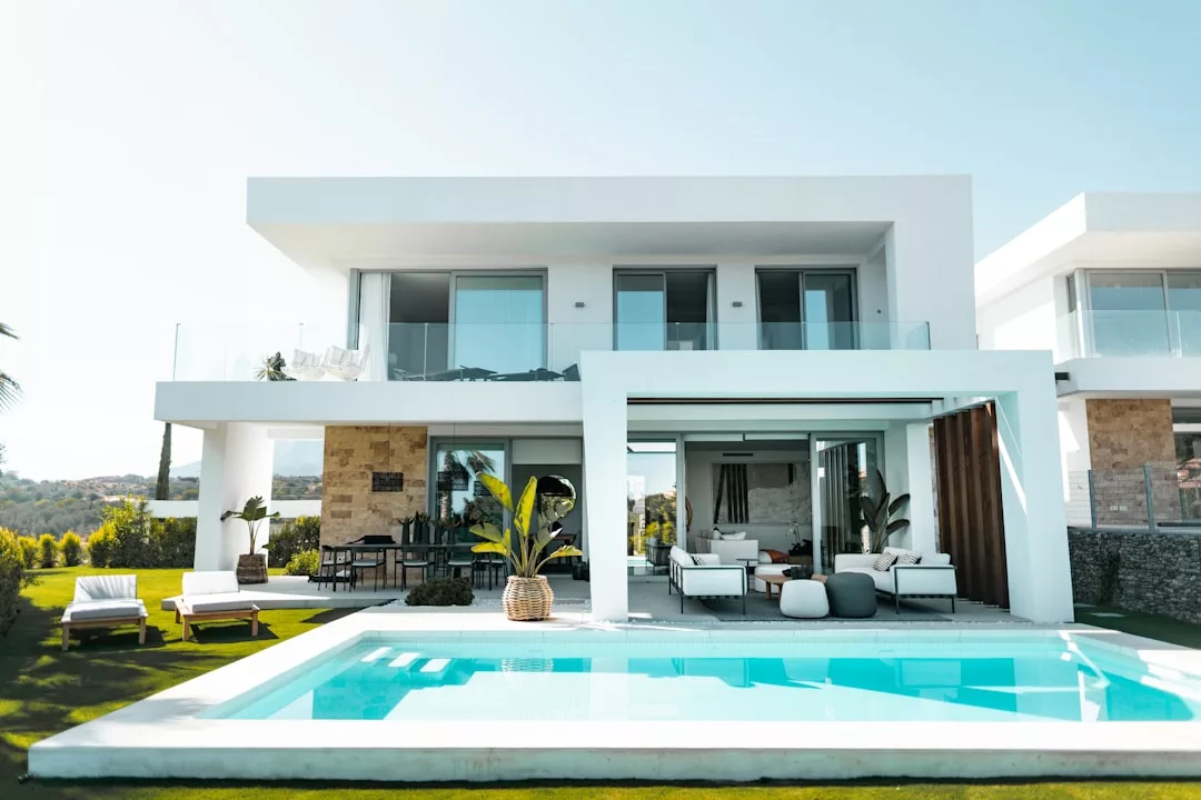 Modern two-story house with large windows, outdoor seating area, swimming pool, and landscaped lawn on a sunny day—an ideal real estate opportunity.