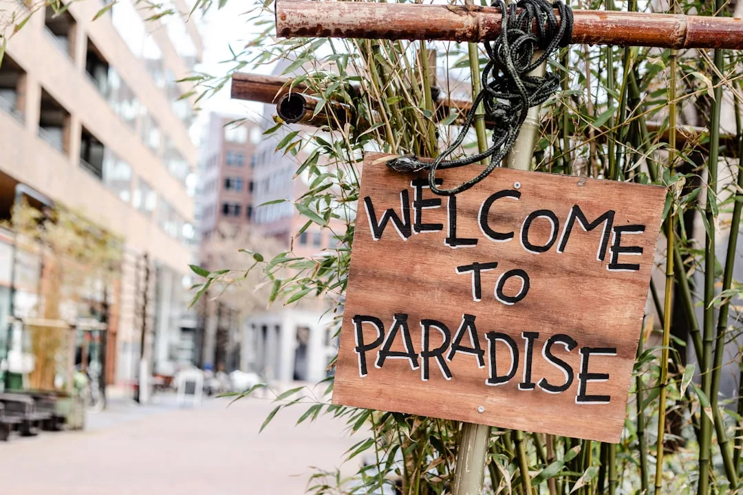 A wooden sign reading "Welcome to Paradise" hangs on bamboo poles, with an urban street and modern real estate buildings in the background.