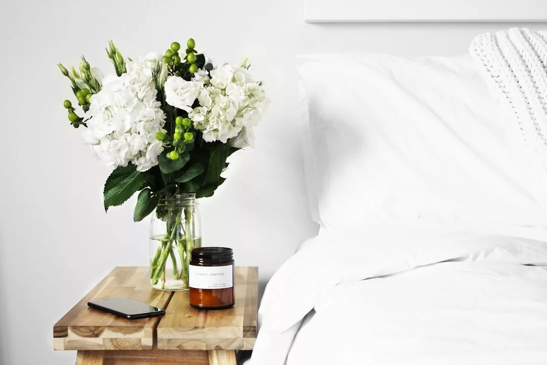 A clear glass vase with white flowers and greenery sits on a wooden bedside table next to a brown candle jar and a smartphone, adding an inviting touch to this Real Estate bedroom with a neatly made white bed.