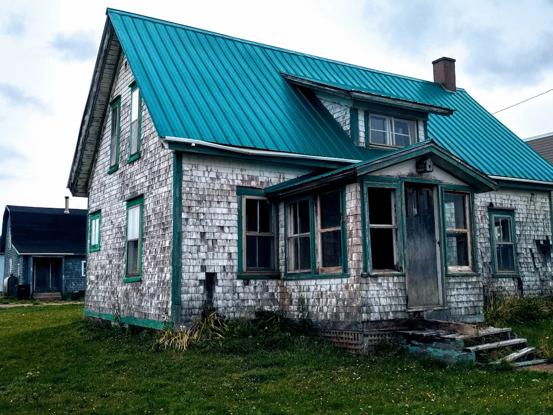 Weathered two-story house with a teal metal roof, peeling paint, and boarded-up windows sits on a grassy lawn under Oslo’s cloudy sky, offering contrast to the city’s focus on sustainable building practices.