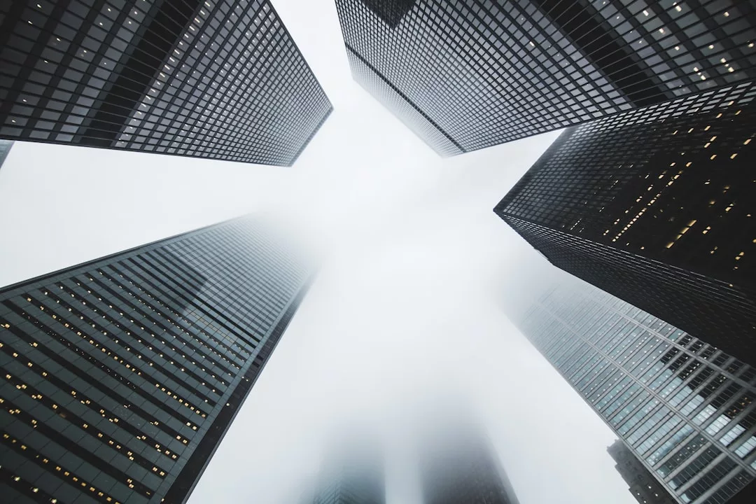 Tall skyscrapers with glass and steel facades, symbols of modern real estate, soar upward and converge into a cloudy, overcast sky as their tops fade into the mist.