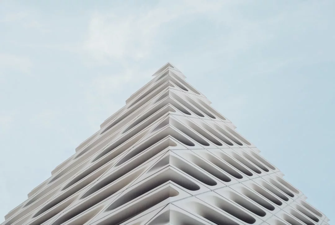 A modern, geometric real estate gem featuring triangular, layered white balconies ascending against a pale blue sky.